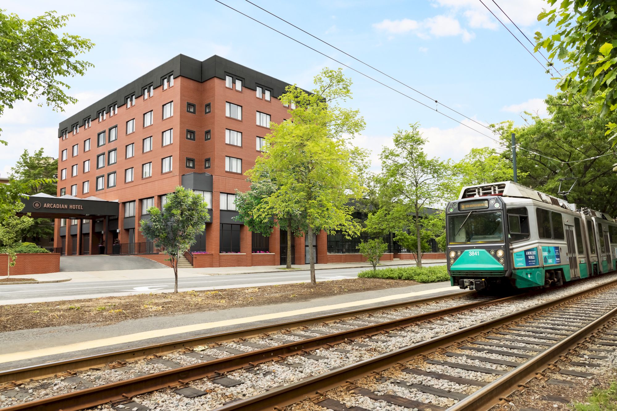 A brick building beside a tram on tracks passes by green trees under a sunny sky, with power lines overhead and a sidewalk nearby.