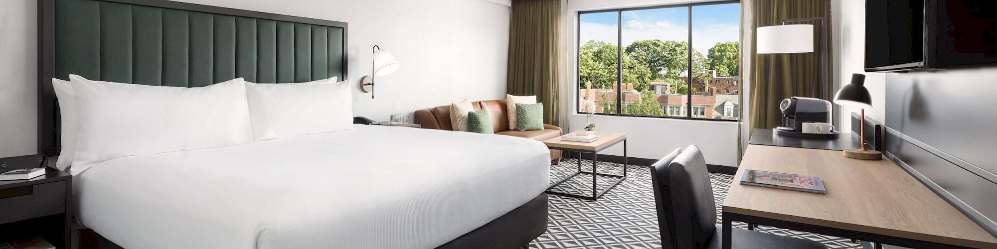 A modern hotel room with a large bed, patterned carpet, desk, chair, and a view of trees through a window.