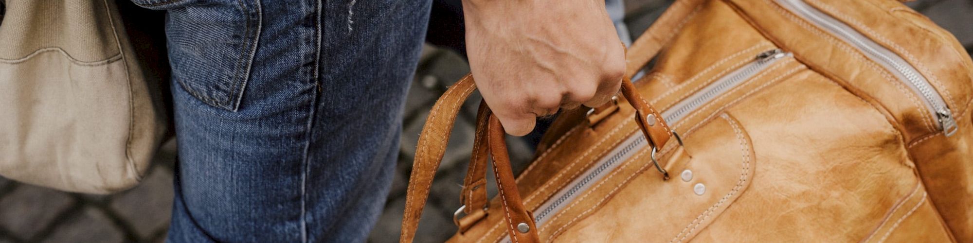 A person in jeans carrying a tan leather duffel bag by its handles, walking on a cobblestone path.