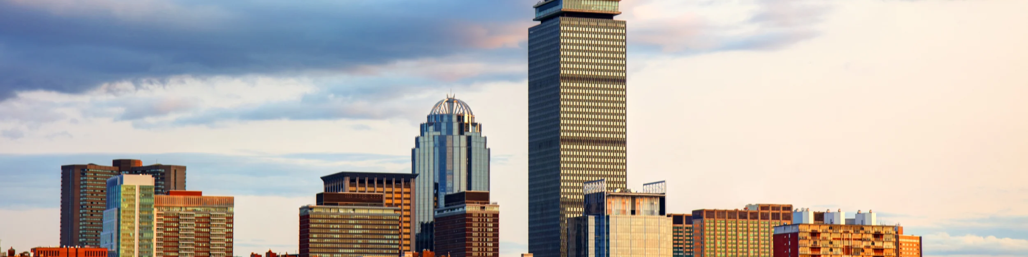 A city skyline with tall office buildings along a river, sailboats on the water, and a partly cloudy sky above.