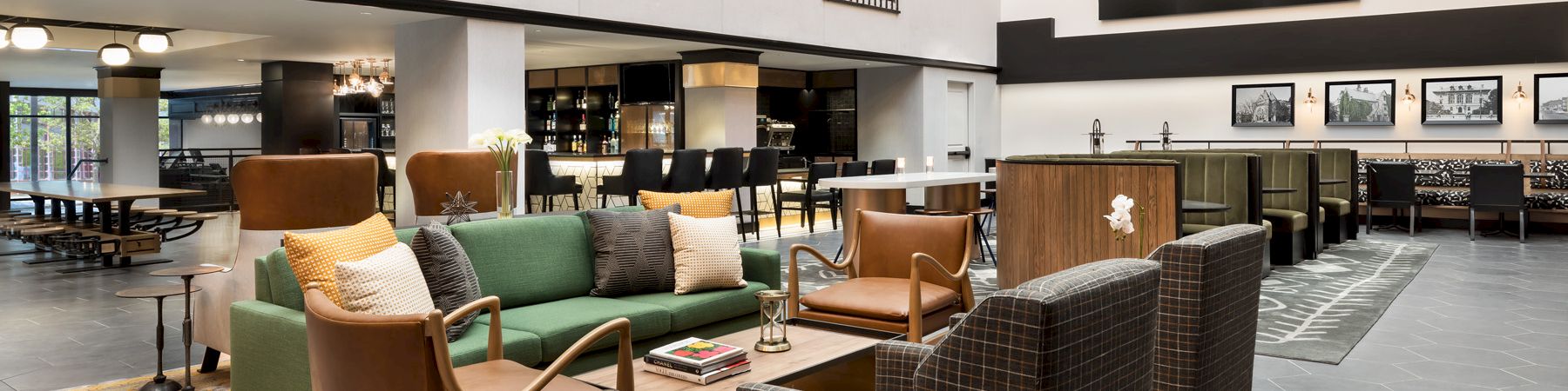 Modern hotel lounge with green and plaid armchairs, coffee tables, a patterned rug, a bar counter, and warm string lights overhead.