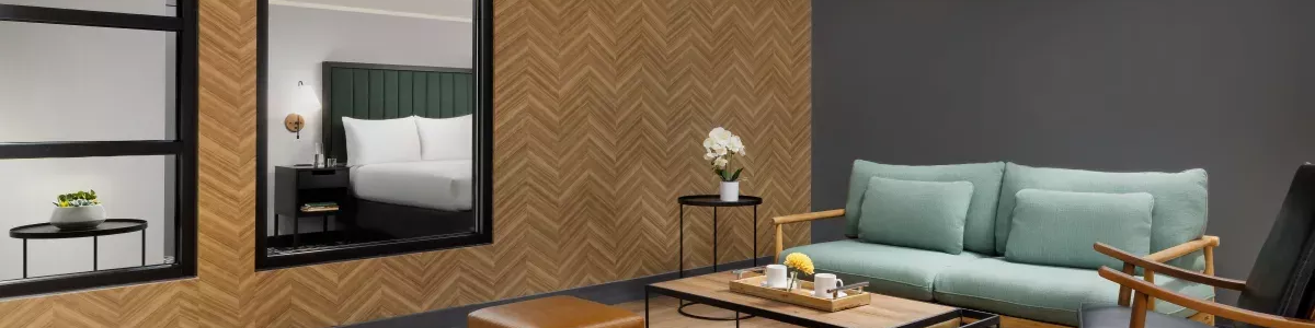 A modern living room with a green couch, wooden rocking chair, two brown ottomans, a glass coffee table, and mustard wall paneling with black trim.