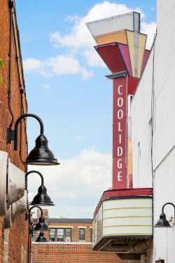 A narrow alley between brick buildings with vintage street lamps, ivy on the left, a red Coolidge sign on the right, and a blue sky above.