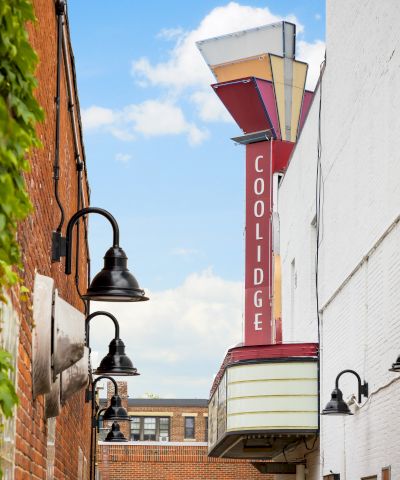 A narrow alley between brick buildings with vintage street lamps, ivy on the left, a red Coolidge sign on the right, and a blue sky above.