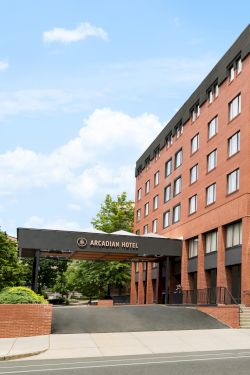 A red brick hotel building with multiple windows, trees lining the sidewalk, and a black-framed entrance canopy at the left.