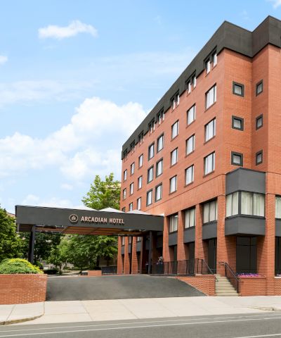 A red brick hotel building with multiple windows, trees lining the sidewalk, and a black-framed entrance canopy at the left.