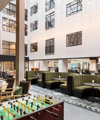 A modern hotel lobby with a foosball table, cozy green couches, glass walls, and a bright open-air feel, inviting guests to relax and socialize.