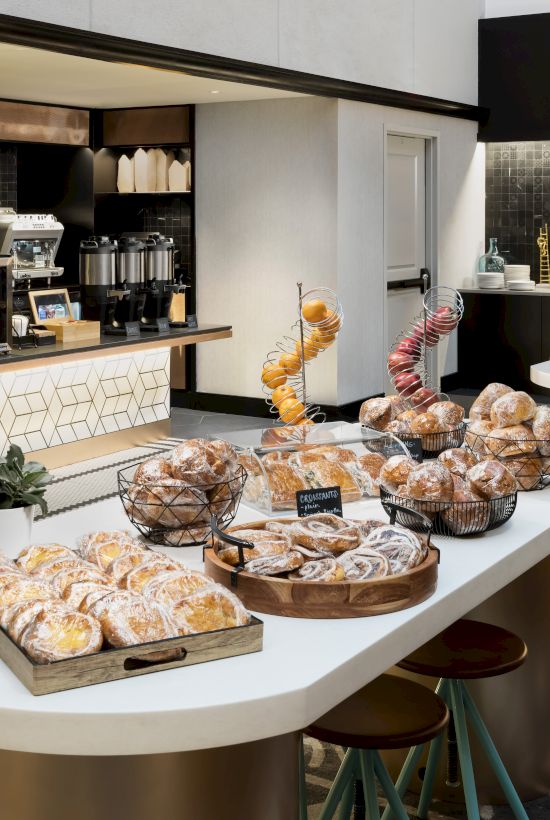 A modern cafe scene with a curved white pastry table, assorted breads and pastries, a coffee station in the back, and seating along a stylish, tiled floor.