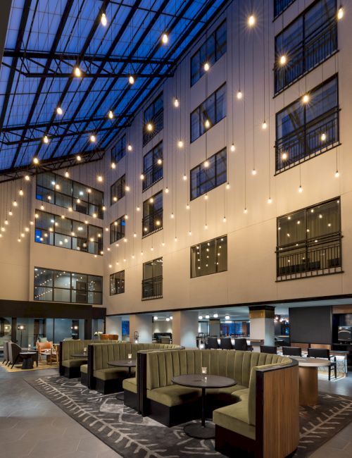 A modern, airy hotel lobby with high ceilings, string lights, and cozy booth seating around round tables.