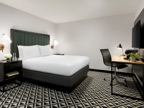 A modern hotel room with a large bed, dark headboard, two wall lights, a desk with a chair, a flat-screen TV, and a black-and-white geometric patterned floor.