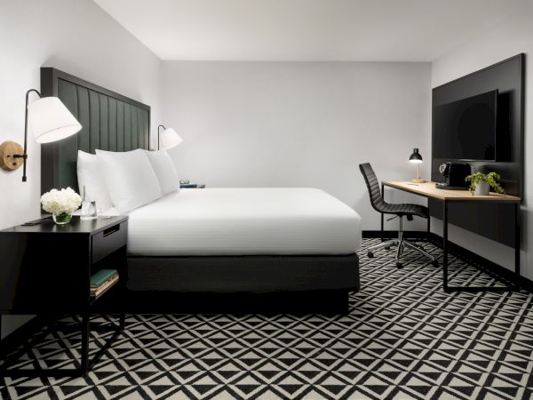 A modern hotel room with a large bed, two black nightstands, a desk and chair, a wall-mounted TV, and a geometric black-and-white patterned carpet.