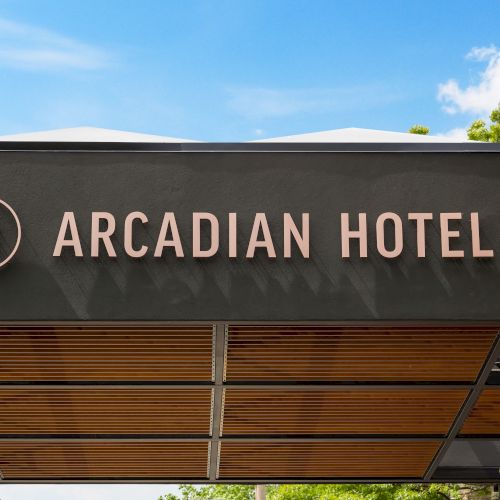 A building sign reads &ldquo;ARCADIAN HOTEL&rdquo; under a blue sky with a few trees and clouds.