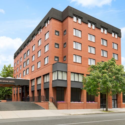 A red-brick mid-rise hotel building labeled &ldquo;Arcadian Hotel,&rdquo; with windows, an entrance canopy, and green trees under a blue sky.