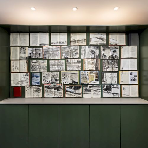 A gallery wall displays dozens of open sketchbooks and black‑and‑white drawings, with small sculptures on side shelves and a green interior.
