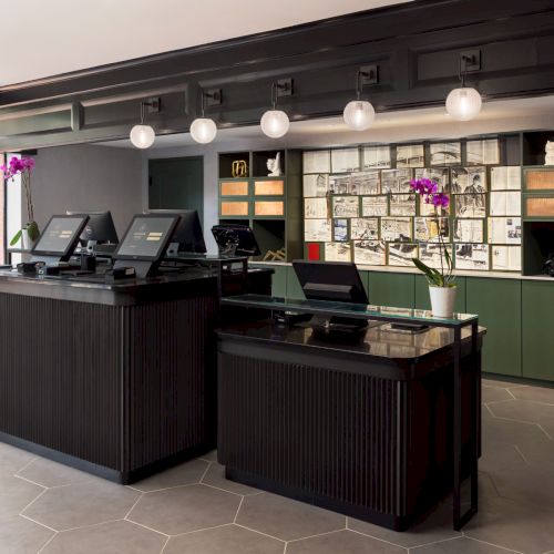 A modern reception area with dark counters, office stations, hanging globe lights, and framed artwork/posters on green shelving.