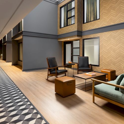 A modern hotel corridor with patterned carpet, wood-paneled walls, and a lounge area featuring chairs, a coffee table, and a sofa.