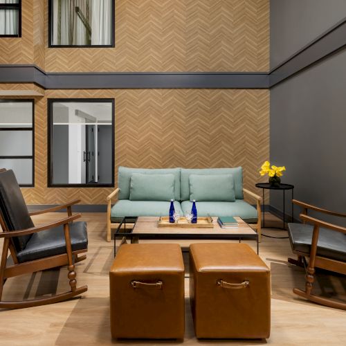 A modern lounge with a teal sofa, leather chairs, ottoman seating, a small table with flowers, and patterned wood-paneled walls.