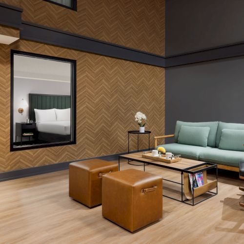 A modern living room with a teal sofa, leather ottoman, coffee table, and a window revealing a neatly styled bedroom.