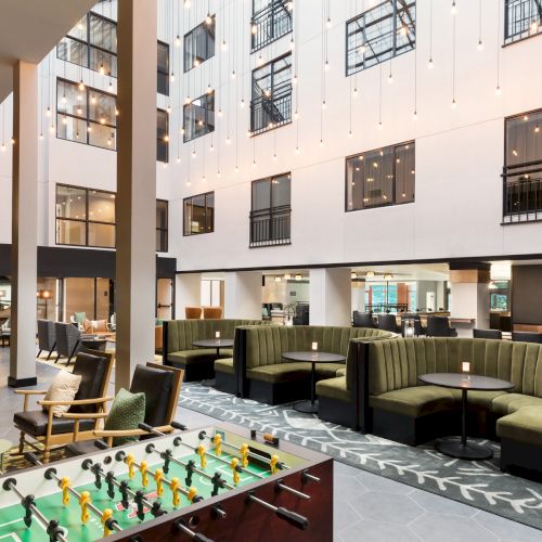 A modern hotel lounge with green sofas, warm hanging string lights, large windows, and a foosball table in the foreground.