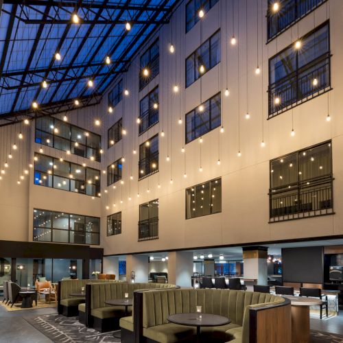 A modern hotel lobby lounge with green booth seating, round tables, large windows, and warm hanging globe lights under a glass ceiling.