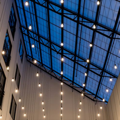 A tall atrium of glass and steel with strings of warm hanging bulbs glowing against the blue sky backdrop.