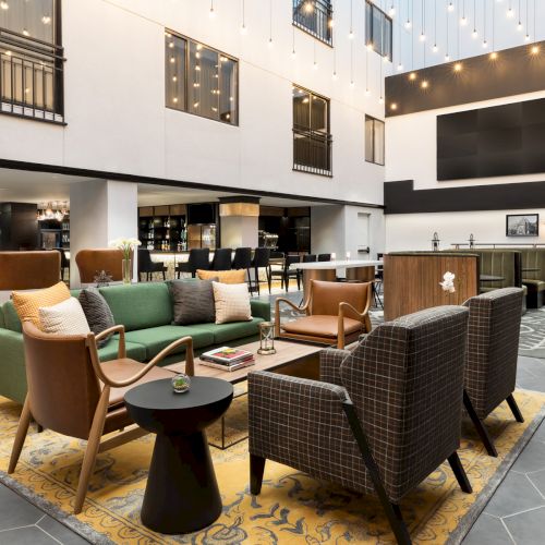 A modern indoor lounge with sofas and chairs, caf&eacute; bar, framed photos, and warm string lights along the high ceiling.
