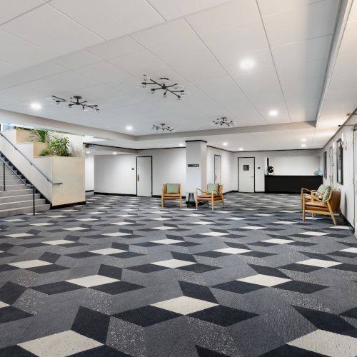 Bright building hallway with doors on the right, carpet tiles, chairs/benches, and ceiling-mounted surveillance drones/cameras overhead.