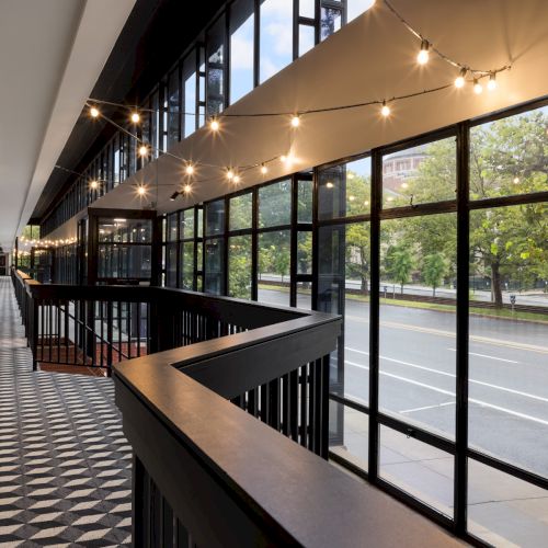 A bright modern building hallway with large windows, string lights, and a railing overlooking a street.