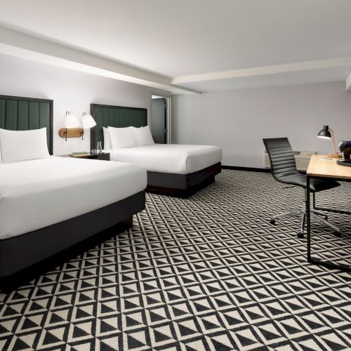 A modern hotel room with twin beds, green headboards, a desk with coffee maker and flowers, a wall TV, and patterned carpet.