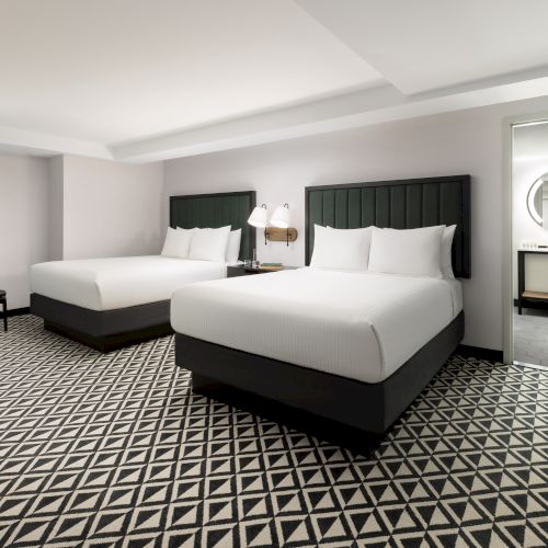 A modern hotel suite with two neatly made beds, dark headboards, patterned carpet, and a bathroom vanity visible through the doorway.