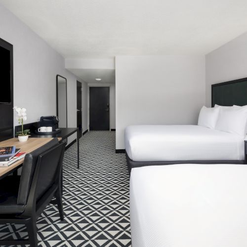 A modern hotel room with a king-size bed, desk with books and coffee setup, wall TV, lamps, and a patterned carpet.