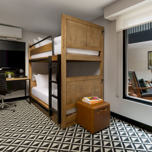 A modern hotel room with a wooden bunk beds, desk setup, chairs by a window, patterned carpet, and a small side cabinet.