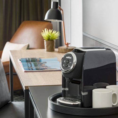 A black Lavazza espresso machine with two white cups sits on a tray in a modern living room, with a sofa, plant, and lamp in view.