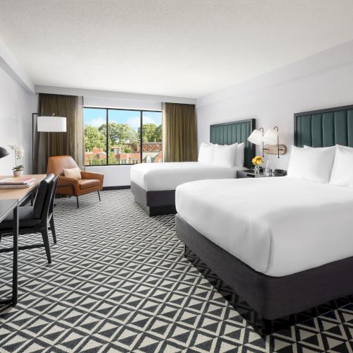 A modern hotel room with large windows, two twin beds with white linens, bedside lamps, a desk, and an armchair.