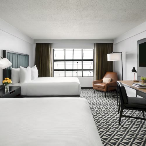 Modern hotel room with two beds, window blinds, desk, chair, floor lamp, and wall-mounted TV.