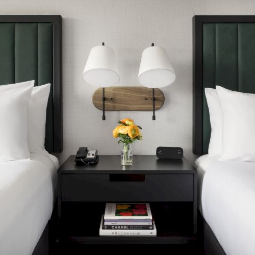 A hotel bedside setup: two beds, a central nightstand with a vase of flowers, and two reading lamps.