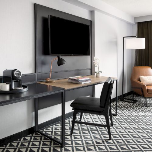 A modern office lounge with a desk, large TV, lamp, coffee machine, chairs, an armchair, and a window with curtains.