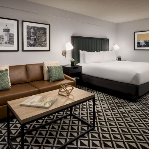 A modern hotel room featuring a sofa, king-size bed, coffee table, patterned carpet, bedside lamps, and framed wall photos.