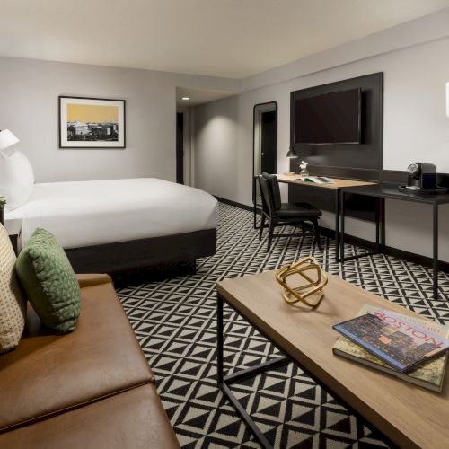 A modern hotel room with a bed, sitting area, patterned carpet, desk setup, and a wall-mounted TV.