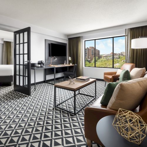 Bright modern hotel suite with sofa, desk, TV, open door, patterned carpet, and city-view window.