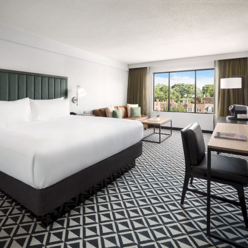 A modern hotel room with a large bed, desk, TV, window view, patterned carpet, and warm lighting.