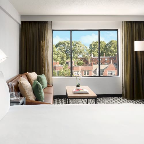 A modern hotel room with a bed, sofa and cushions, a coffee table, tall curtains, and a large window showing trees and buildings.