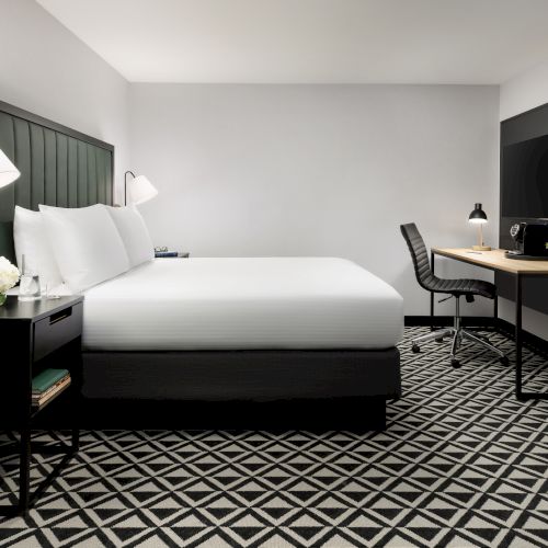 A modern hotel room with a large bed, desk and chair, TVs, lamps, and a patterned carpet.