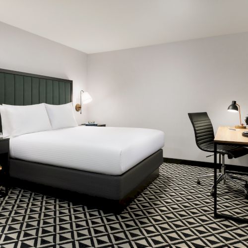 A modern hotel room: a bed with pillows, bedside tables, a desk with chair and lamp, and a wall TV.