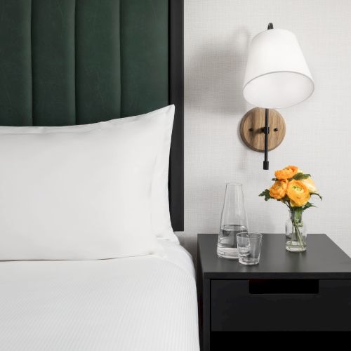 A modern bedroom scene with a bed, a bedside table, a wall-mounted lamp, orange roses in a vase, and drinking glasses.