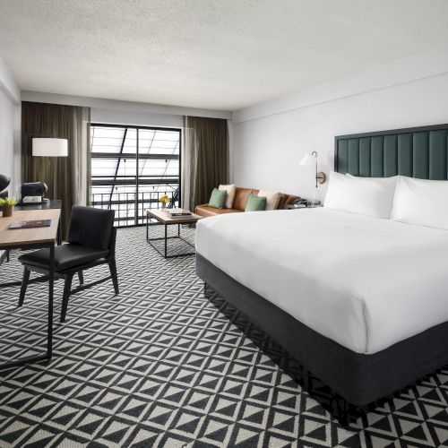A modern hotel room with a large bed, desk, lamps, and a window with curtains, lit by daylight.