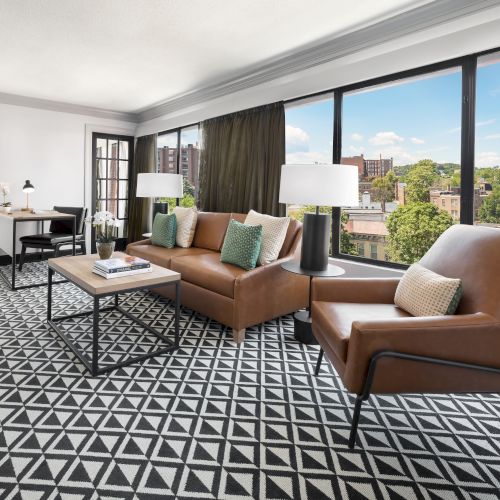 A modern living room with brown leather sofas, a patterned carpet, a coffee table, and tall windows overlooking a city skyline.