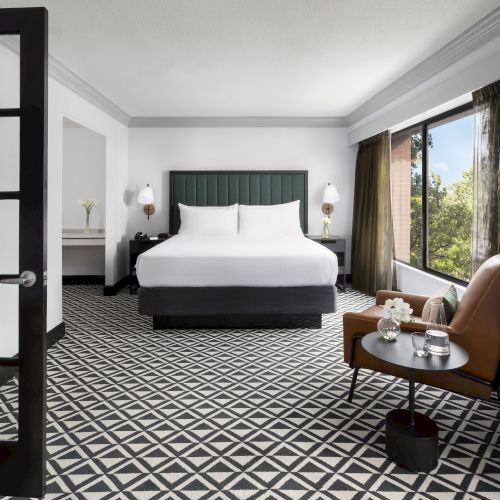 A modern hotel bedroom with a large bed, bedside lamps, patterned carpet, and a seating table by big windows with curtains.