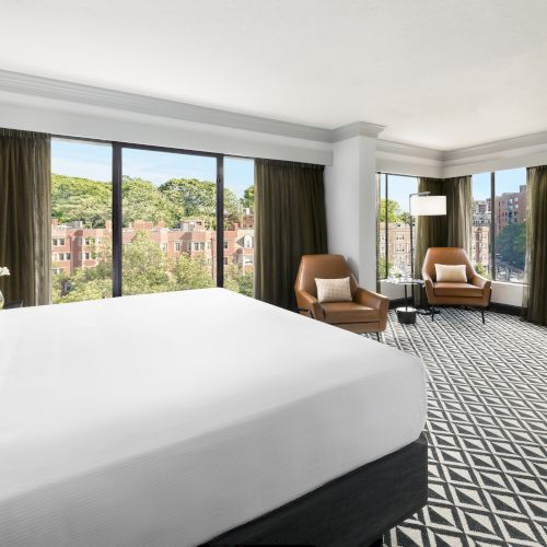 A bright hotel room with a large bed, lounge chairs, dark curtains, and windows overlooking city greenery.