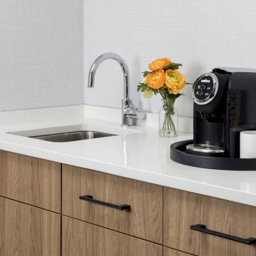 A modern kitchen sink with a chromed faucet, a black coffee machine, white mugs, and a vase of orange-yellow roses on the counter.